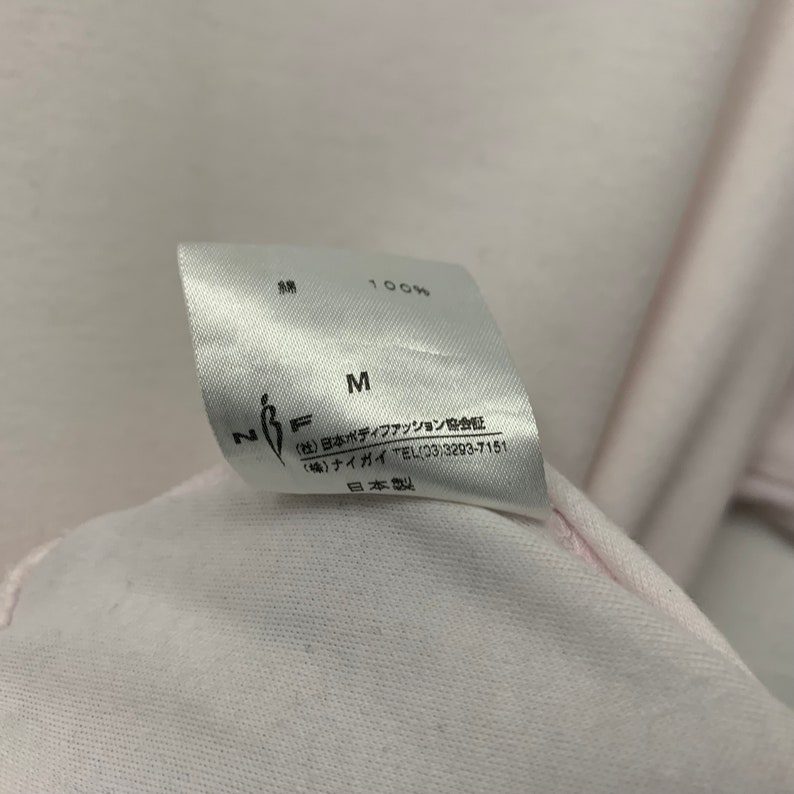 May include: Close-up of a white clothing tag with Japanese text and the letters "M" and "N F". The tag indicates 100% cotton. The text includes the name of a Japanese body fashion association and a phone number.