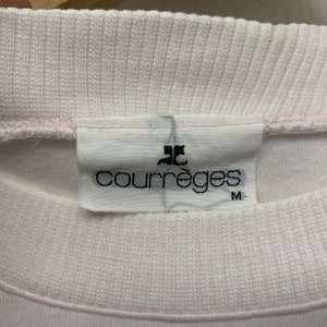 May include: Close-up of a white sweatshirt collar with a tag. The tag features the brand name "Courr&egrave;ges" in black font, along with a logo and the size "M". The sweatshirt appears to be made of a soft, textured fabric.