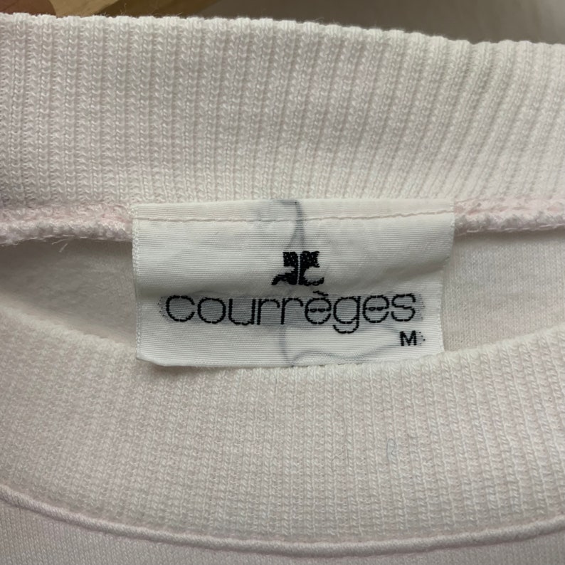 May include: Close-up of a white sweatshirt collar with a tag. The tag features the brand name "Courr&egrave;ges" in black font, along with a logo and the size "M". The sweatshirt appears to be made of a soft, textured fabric.