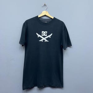 May include: Black t-shirt with a white DC logo and crossed skateboards. The logo is centered on the front of the shirt. The shirt is hanging on a wooden hanger against a gray background.