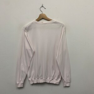 May include: A long-sleeved, white sweatshirt hangs from a wooden hanger against a white wall. The sweatshirt has a crew neck and ribbed cuffs and hem. The fabric appears to be a soft, lightweight material.