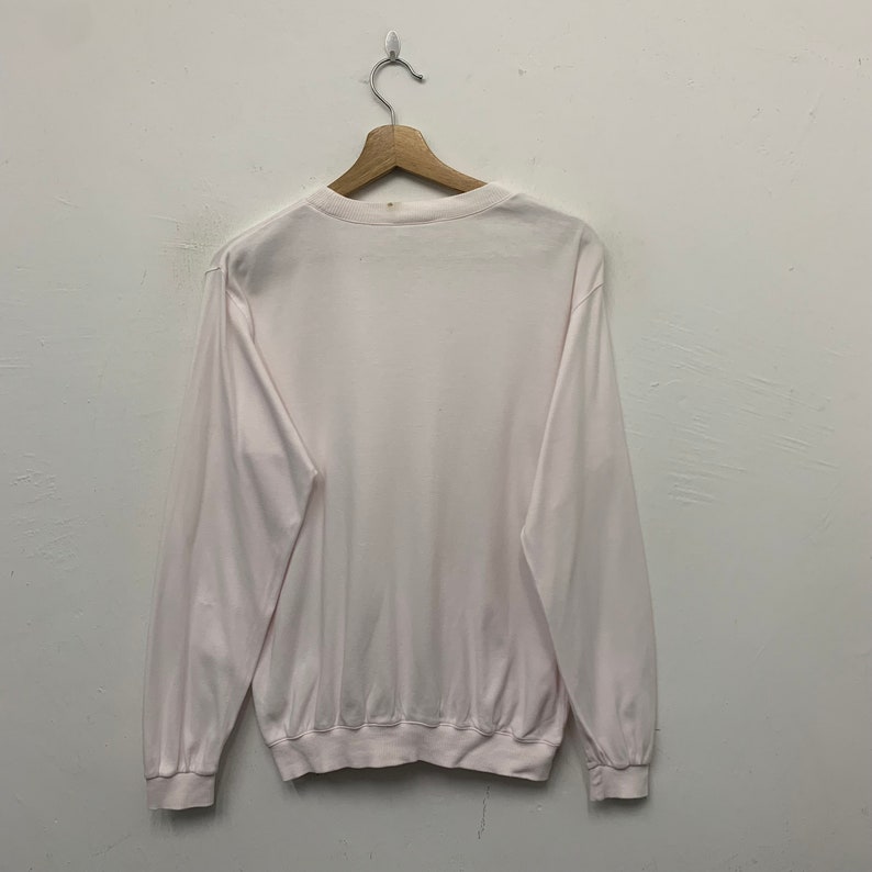 May include: A long-sleeved, white sweatshirt hangs from a wooden hanger against a white wall. The sweatshirt has a crew neck and ribbed cuffs and hem. The fabric appears to be a soft, lightweight material.