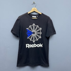 Op de afbeelding: Zwart Reebok T-shirt met een wit en blauw sterrenpatroon op de voorkant. Het Reebok-logo staat eronder in het wit. Het T-shirt hangt aan een houten hanger. Gemaakt van zacht materiaal.