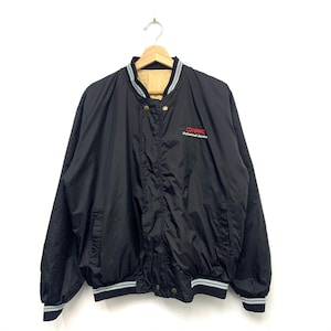 May include: Black nylon bomber jacket with a beige lining, featuring the text "COMPAQ Professional Service" in red. The jacket has a button-up front, a striped collar, and cuffs. The jacket is hanging on a wooden hanger.