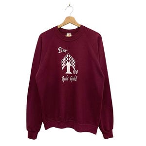 May include: A maroon sweatshirt with a white graphic of a tree and the text "Pine Tree Quilt Guild". The long-sleeved top is hanging on a wooden hanger. The sweatshirt has a crew neck and ribbed cuffs.
