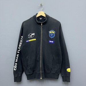 May include: Black zip-up jacket with multiple patches and text. The jacket features a "BRASIL" patch with a blue background and stars, and the text "CEU MAR FLORESTA" down the left sleeve. The jacket has a stand-up collar and two front pockets.