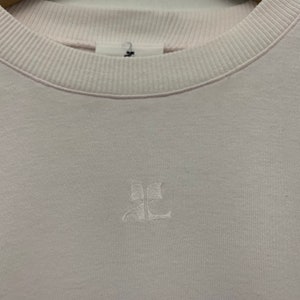 May include: A light pink sweatshirt with a ribbed crew neck and cuffs. The sweatshirt features a small, embroidered logo on the chest. The fabric appears to be a soft, comfortable material.