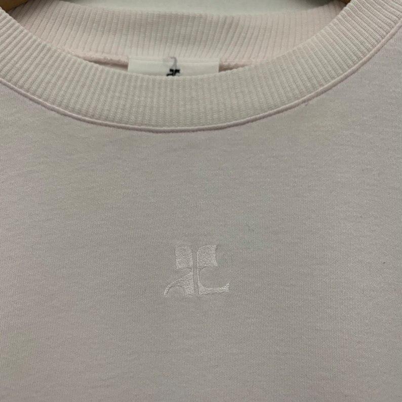 May include: A light pink sweatshirt with a ribbed crew neck and cuffs. The sweatshirt features a small, embroidered logo on the chest. The fabric appears to be a soft, comfortable material.