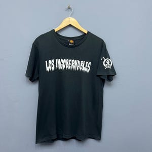 Peut inclure: T-shirt noir avec l'inscription blanche "LOS INGOBERNABLES" sur la poitrine. Un petit motif est présent sur la manche droite. Le t-shirt est suspendu à un cintre en bois.