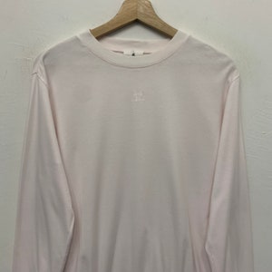 May include: Pale pink long-sleeve shirt with a crew neck. The shirt has a small, embroidered design on the chest. The shirt is hanging on a wooden hanger against a white background. The shirt is made of a soft material.