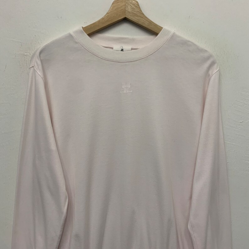 May include: Pale pink long-sleeve shirt with a crew neck. The shirt has a small, embroidered design on the chest. The shirt is hanging on a wooden hanger against a white background. The shirt is made of a soft material.