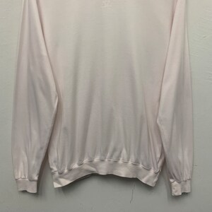 May include: A pale pink long-sleeved sweatshirt with a ribbed hem and cuffs. The crew neck sweatshirt has a small logo on the upper back. The fabric appears soft and lightweight, suitable for casual wear.