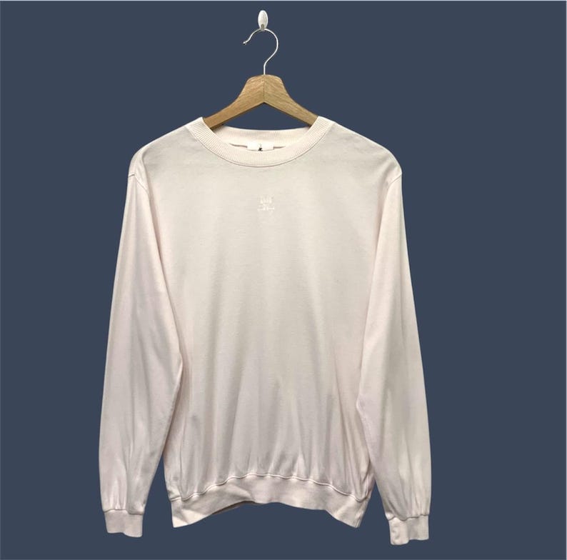 May include: A light pink long-sleeved shirt hanging on a wooden hanger. The shirt has a crew neck and a small, embroidered logo on the chest. The sleeves are cuffed at the wrists, and the bottom hem is ribbed.