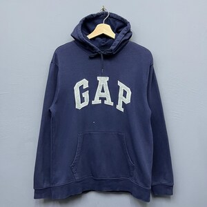Könnte beinhalten: Marineblauer Kapuzenpullover mit dem Wort "GAP" in weißen Blockbuchstaben auf der Brust. Der Pullover hat eine Vordertasche und eine Kapuze mit Kordelzug. Das Kleidungsstück hängt an einem Holzbügel.