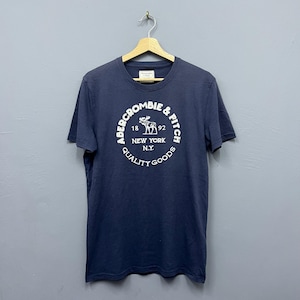 May include: Navy blue Abercrombie & Fitch t-shirt with a white circular logo. The logo features the brand name, a deer graphic, and the text "New York N.Y. Quality Goods". The shirt is hanging on a wooden hanger.