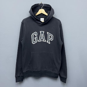 May include: Black hooded sweatshirt featuring the word "GAP" in white with a black outline on the chest. The sweatshirt has a front pocket and a drawstring hood. The garment is suspended on a wooden hanger.