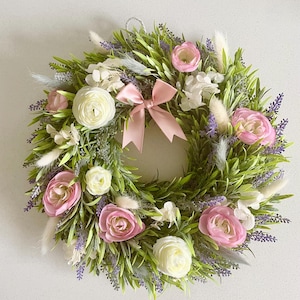 Könnte beinhalten: Ein dekorativer, kreisförmiger Kranz mit künstlichen Blumen und Grünzeug. Er zeigt rosa Rosen, weiße Hahnenfußblüten und Lavendel, mit einer hellrosa Schleife. Die Basis besteht aus grünen Blättern und weißen Akzenten.