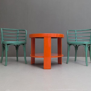 Könnte beinhalten: Ein Möbelset im Mid-Century-Modern-Stil. Es besteht aus einem runden, orangefarbenen Tisch mit einem unteren Regal und zwei grünen Stühlen. Die Stühle haben eine geschwungene Rückenlehne mit horizontalen Lamellen. Das Set steht vor grauem Hintergrund.