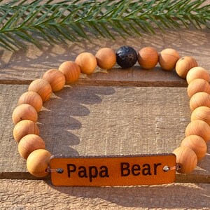 Puede incluir: Un brazalete de cuentas de madera con una banda de cuero marrón que dice "Papa Bear". El brazalete tiene una cuenta de lava negra.