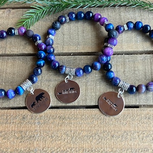 May include: Three blue and purple tiger's eye beaded bracelets with silver charms. The charms have a bear and arrow design and the text "Mama Bear" and "Papa Bear".