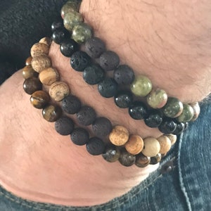 May include: Three beaded bracelets stacked on a person's wrist. The top bracelet is green and black, the middle bracelet is black, and the bottom bracelet is brown and black.