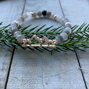 May include: A beaded bracelet featuring gray and white stone beads with a rose gold mountain range charm. The bracelet is displayed on a wooden surface with green sprigs.