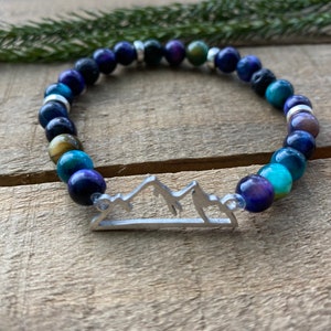 Pode incluir: Um bracelete de contas azuis, verdes e douradas com um amuleto de montanha prateado. O bracelete é feito de contas de olho de tigre, lápis-lazúli e turquesa.