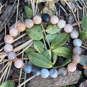 May include: A beaded bracelet featuring a mix of round, polished gemstones in shades of peach, gray, and white. The bracelet includes a black lava bead and is accented with rose gold-colored spacers. The bracelet is displayed on a bed of leaves and wood chips.