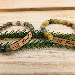 Può includere: Due braccialetti di perline con targhette di legno con la scritta "Let Them". Un braccialetto presenta perline grigie e rosa, l'altro ha perline gialle, blu e marroni. Entrambi sono esposti su verde su uno sfondo di legno chiaro.