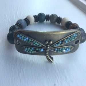 Könnte beinhalten: Ein Armband mit Perlen und einem Libellen-Anhänger. Das Armband besteht aus grünen, braunen und weißen Perlen. Der Libellen-Anhänger ist aus Metall und hat blaue und grüne Akzente.