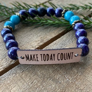 May include: A blue and turquoise beaded bracelet with a brown leather tag that reads "MAKE TODAY COUNT".
