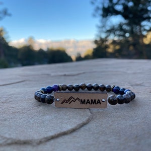 May include: A black and brown beaded bracelet with a leather tag that says "MAMA" and a mountain range design.