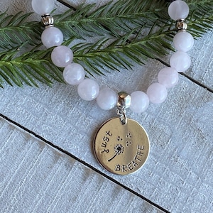 May include: A pink rose quartz beaded bracelet with a silver charm that says "Just Breathe" with a dandelion design.