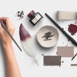 Könnte beinhalten: Draufsicht auf Kalligraphie-Zubehör, darunter eine schwarze Tintenflasche mit burgunderfarbenem Deckel, ein schwarzer Stift und eine kleine Schale mit Federn. Eine Feder mit burgunderfarbenen und weißen Farbtönen und ein Wachssiegel sind ebenfalls vorhanden.