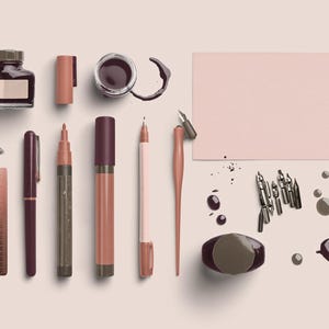 May include: An assortment of stationery items in shades of brown and pink, including ink bottles, pens, markers, a ruler, and a blank card. The items are arranged on a light pink surface, with ink splatters and nibs scattered around.