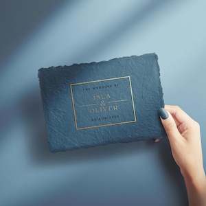 Puede incluir: Una invitación de boda azul oscuro texturizada con letras doradas y un borde dorado. El texto dice "The Wedding of Isla & Oliver 05 Ago 2025". La invitación es sostenida por una mano con esmalte de uñas azul.