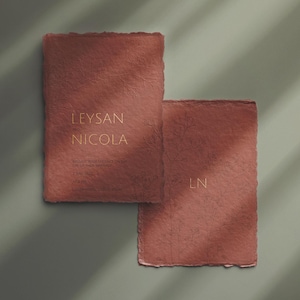 May include: Two textured, reddish-brown paper cards with torn edges. The top card features the names "LEYSAN NICOLA" in gold lettering. The bottom card has the initials "LN" in gold. The cards are on a muted green background.