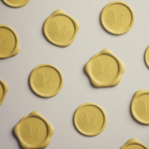 May include: Mustard yellow wax seals with a botanical design and the letter "M". The seals are arranged on a light gray surface. The seals have a circular shape with a decorative border.