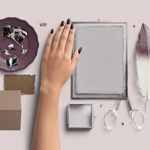 Könnte beinhalten: Draufsicht-Komposition mit einem lila Teller, metallischen Elementen, einem gerahmten Papier, einer Feder und einer Schere. Eine Hand mit dunklem Nagellack ist ebenfalls vorhanden, zusammen mit einer kleinen Box und einer Papierprobe.