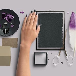 May include: A still-life composition featuring a black plate with purple foil, a framed dark paper, a decorative feather with purple and white hues, and a pair of scissors. A hand with black nail polish is also present.