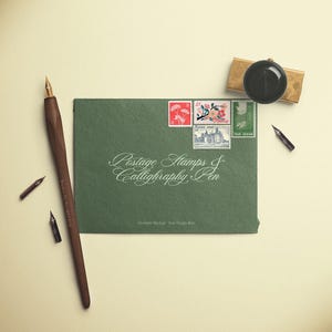 May include: An overhead shot features a dark green envelope with postage stamps and the text "Postage Stamps & Calligraphy Pen." A wooden calligraphy pen, ink nibs, and a stamp holder are also present. The items are arranged on a cream-colored surface.