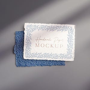 Puede incluir: Una maqueta de papel hecha a mano con un borde rasgado. El papel superior es blanco con un borde floral azul y el texto "Handmade Paper MOCKUP". Debajo hay un papel azul con un patrón floral similar.
