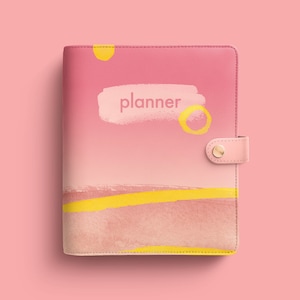 Puede incluir: Una agenda de cuero rosa con un diseño abstracto amarillo y rosa. La palabra "planner" está escrita en blanco en la portada.