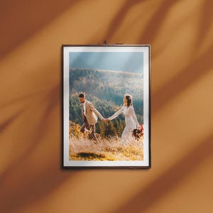Könnte beinhalten: Gerahmtes Foto eines Paares, das sich an den Händen haltend durch ein Feld bewegt. Der Mann trägt einen beigen Anzug, die Frau ein weißes Brautkleid. Der Hintergrund zeigt einen Wald und Sonnenlicht.