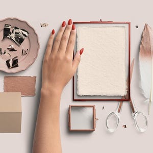 May include: A flat lay featuring a hand with red nails, a pink plate with gold foil, a framed paper, a small square frame, scissors, a feather, and a folded card. The items are arranged on a light pink surface.