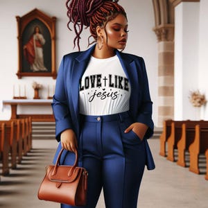 Könnte beinhalten: Eine Frau in einem marineblauen Hosenanzug und einem weißen T-Shirt mit dem Text "LOVE + LIKE Jesus". Sie hält eine braune Lederhandtasche. Der Hintergrund ist ein Kircheninnenraum mit Holzbänken und einem Gemälde.