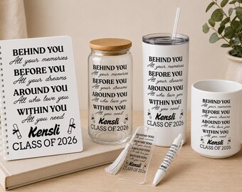 May include: A collection of graduation-themed gifts, including a spiral-bound notebook, a glass jar with a wooden lid, a tumbler with a straw, a coffee mug, a bookmark, and a pen. Each item features the text "BEHIND YOU...CLASS OF 2026".