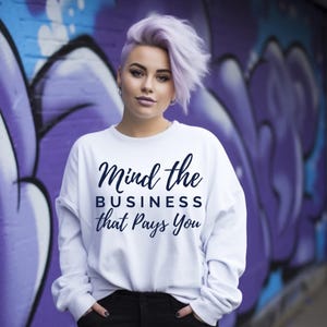 Könnte beinhalten: Ein weißes Langarm-Sweatshirt mit dem Text "Mind the BUSINESS that Pays You" in dunkelblauer Schrift. Die Person, die das Sweatshirt trägt, hat kurze, lavendelfarbene Haare und steht vor einer lila Graffiti-Wand.