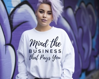 May include: A white long-sleeve sweatshirt with the text "Mind the BUSINESS that Pays You" in a dark blue script. The sweatshirt is worn by a person with short, lavender hair, standing in front of a purple graffiti wall.