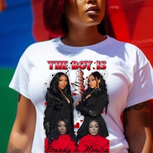 Pode incluir: Camiseta branca com um design gráfico. O texto "THE BOY IS" está acima de imagens de duas mulheres. Os nomes "Brandy & Monica" são impressos abaixo. O fundo é um mural colorido.